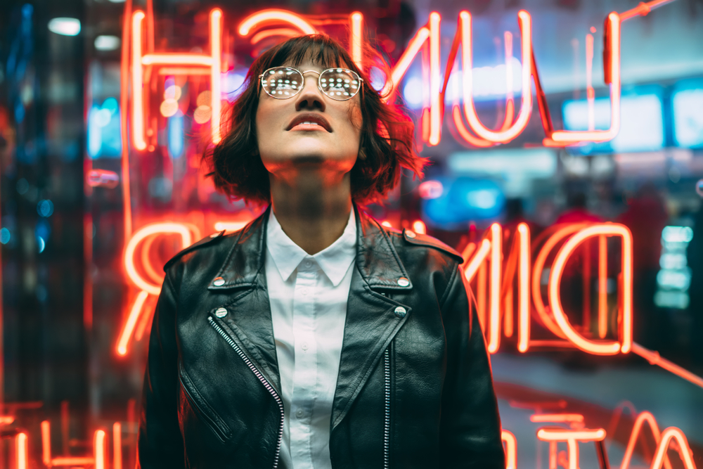 A woman looks up at a sign in New York City: she knows exactly what to wear in New York in fall with her chic white button down, her black leather jacket and her round, gold glasses. The neon sign of the dinner in front of her reflects behind her in hues of red. 