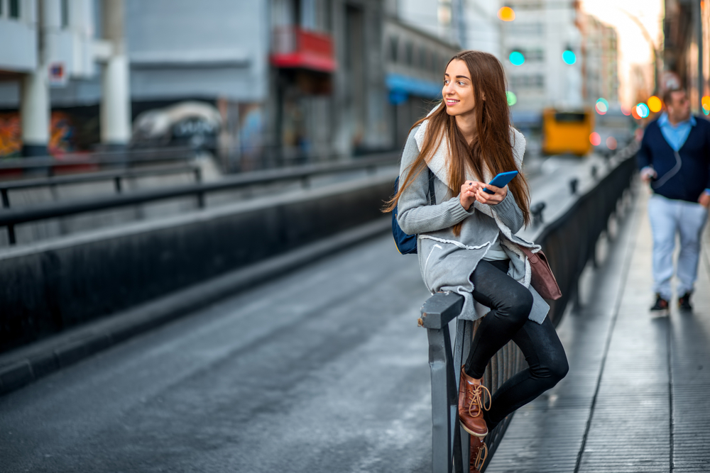 A young girl perches on the edge of a railing waiting for public transport: she wars faux leather leggings, boots and a coat. 