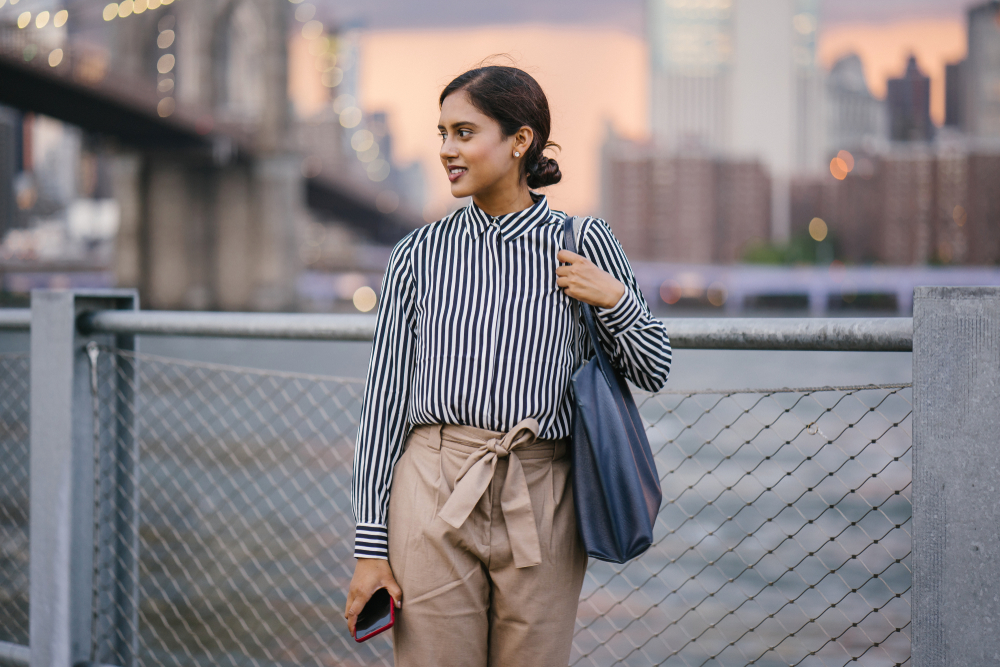 A woman stands on a bridge in New York, the city behind her, in a striped blouse and high waisted pants. She carries a blue tote on her shoulder. 