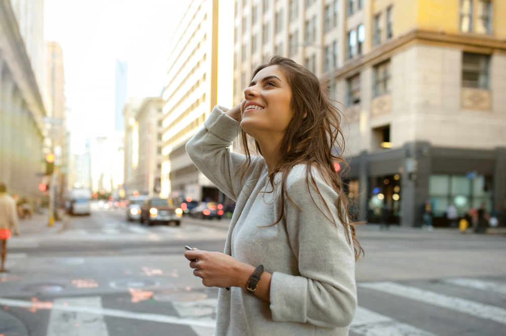 A woman looks up at the buildings in New York, running her hand through her hair, comfortable in her sweater as it is soft and perfect for an option when wondering what to wear in New York in fall. 