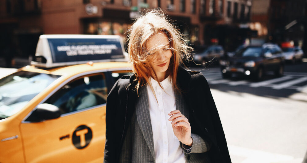 woman standing in front of a yellow taxi wearing a long black coat and white undershirt