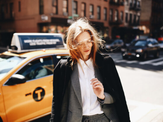 woman standing in front of a yellow taxi wearing a long black coat and white undershirt