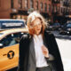 woman standing in front of a yellow taxi wearing a long black coat and white undershirt