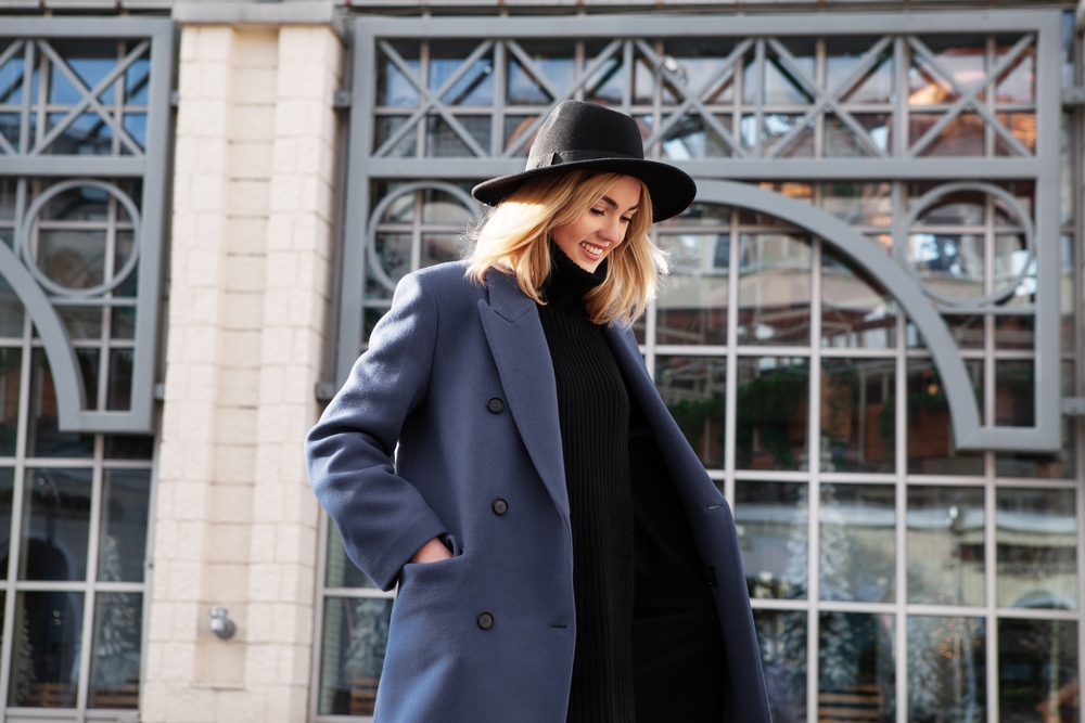 A woman walks by Grand Central Statin wearing a black turtle neck dress and a wool, blue coat over it. She pairs it with a nice black hat. 