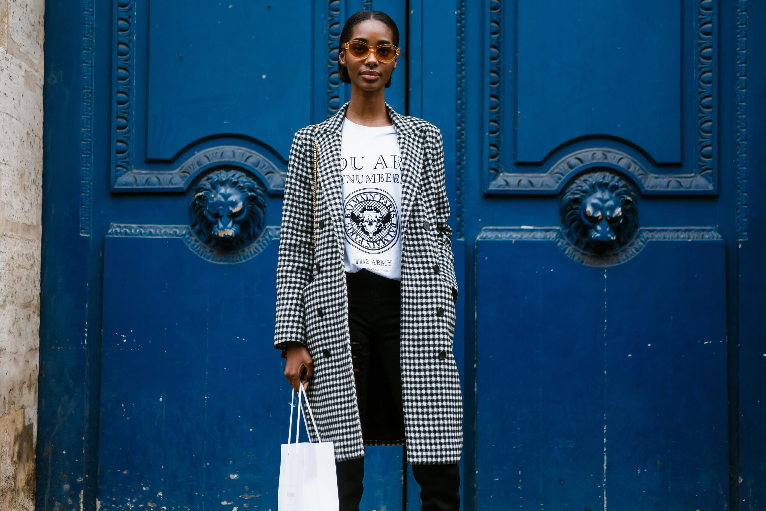 A woman in a plaid jacket, skirt, knee high boots and a graphic tee stands in front of a blue door, showing us exactly what to wear in Paris in fall. 