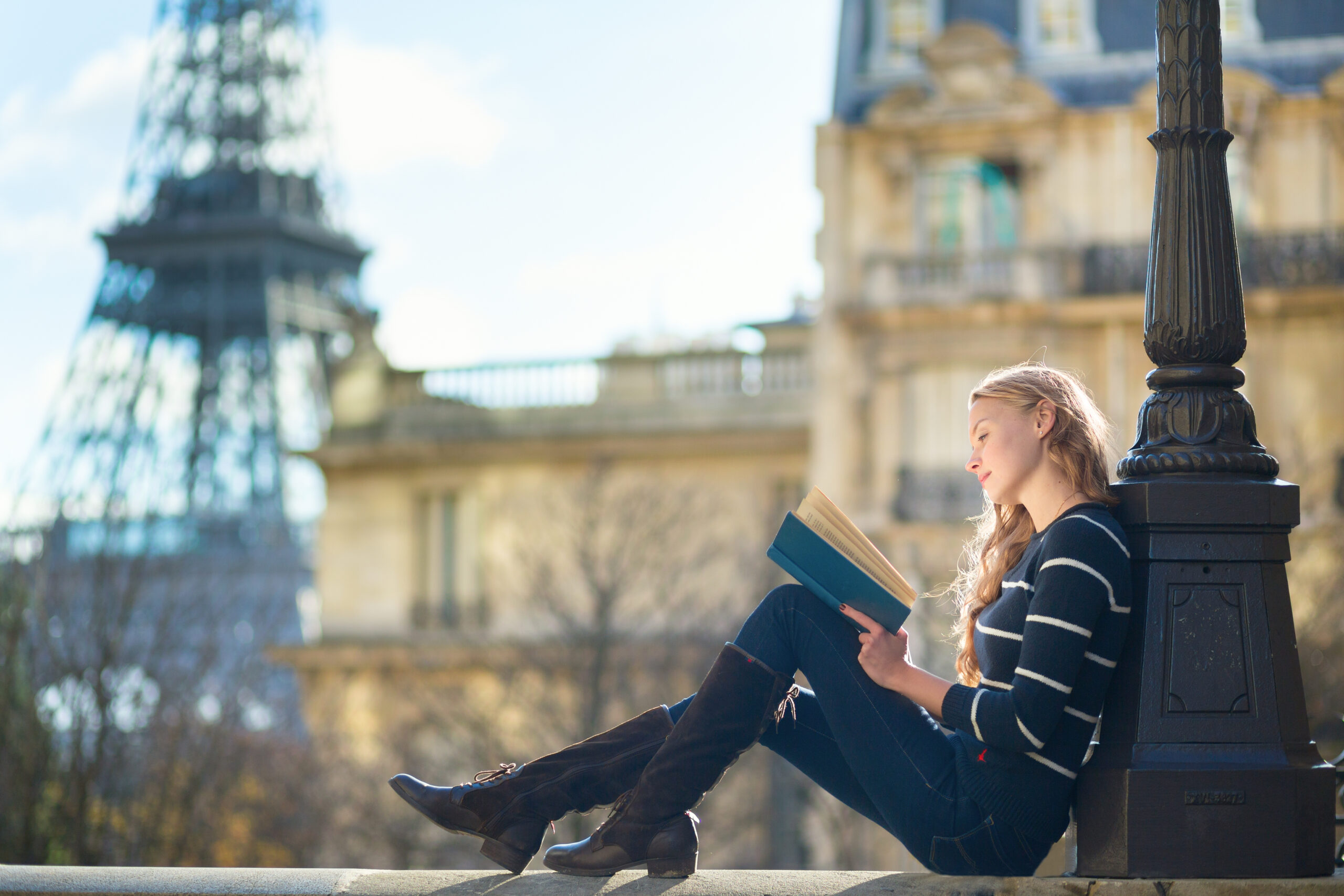 A woman sits on the edge of the city in jeans and a sweater, reading a book with the Eiffel Tower behind her. 