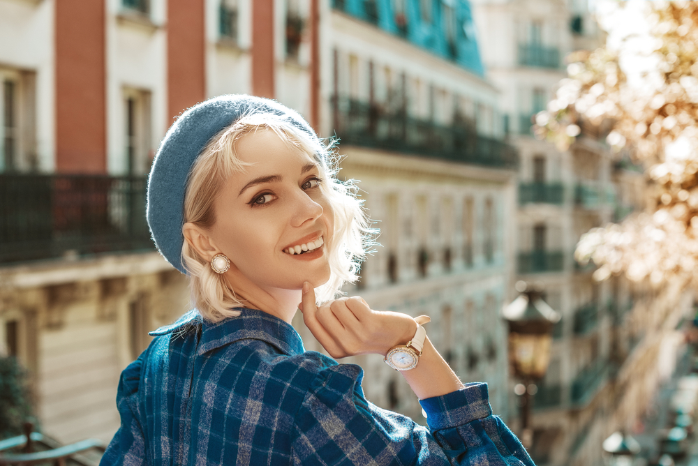 A blonde woman looks over her shoulder in a plaid shirt and blue beret, wearing a matching watch and earrings. 