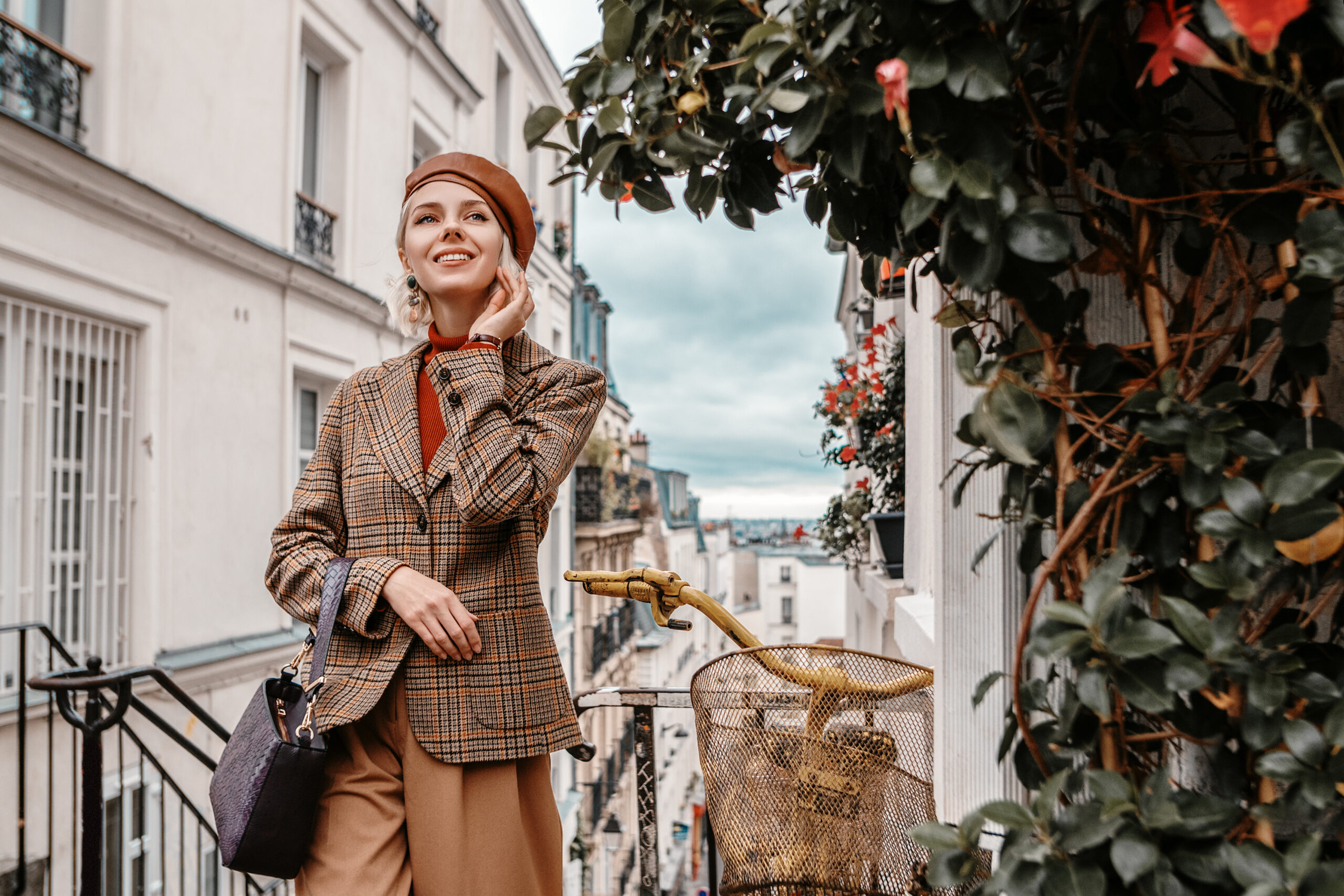 While going up the stairs, a woman wears trousers, a plaid coat, and a turtle neck that matches the color schemes of the Paris buildings behind her. 