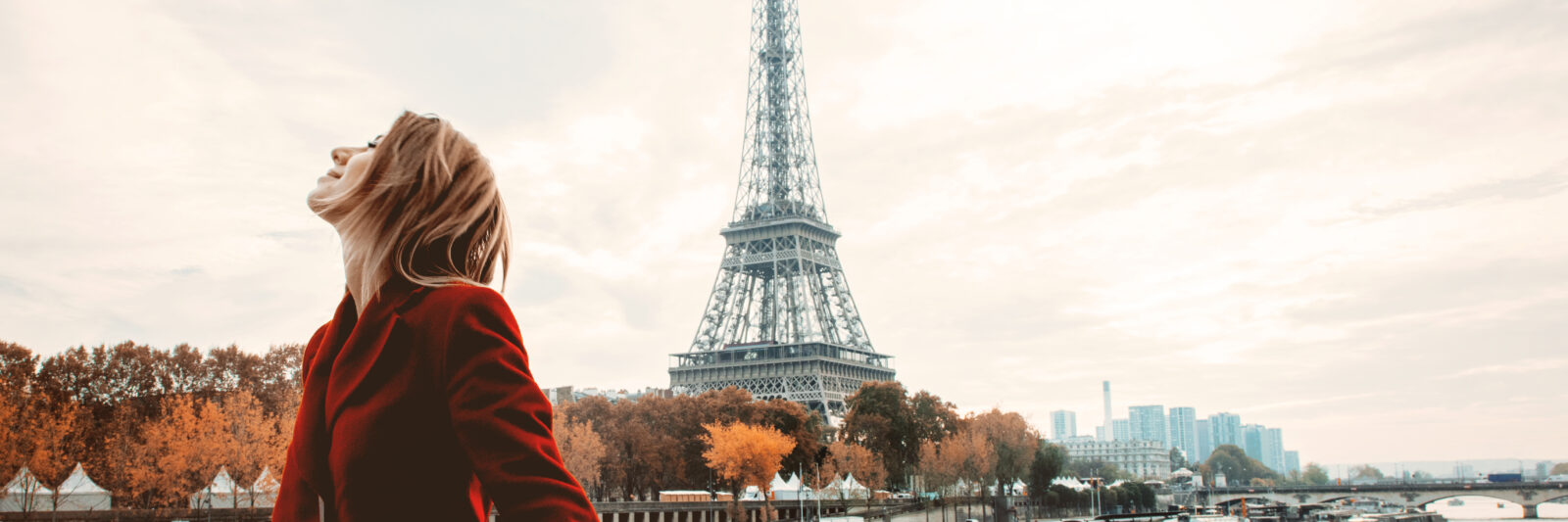 A woman in a red jacket knows exactly what to wear in Paris in the fall: she dawns a wool red jacket and is comfortable as she stands on the Siene walkway, the Eiffel Tower behind her.