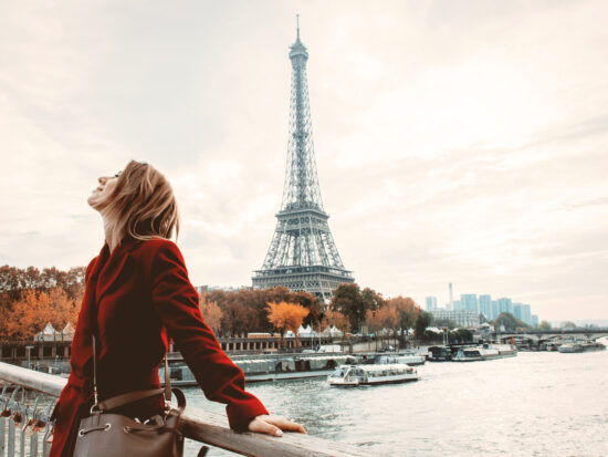 A woman in a red jacket knows exactly what to wear in Paris in the fall: she dawns a wool red jacket and is comfortable as she stands on the Siene walkway, the Eiffel Tower behind her.