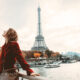 A woman in a red jacket knows exactly what to wear in Paris in the fall: she dawns a wool red jacket and is comfortable as she stands on the Siene walkway, the Eiffel Tower behind her.