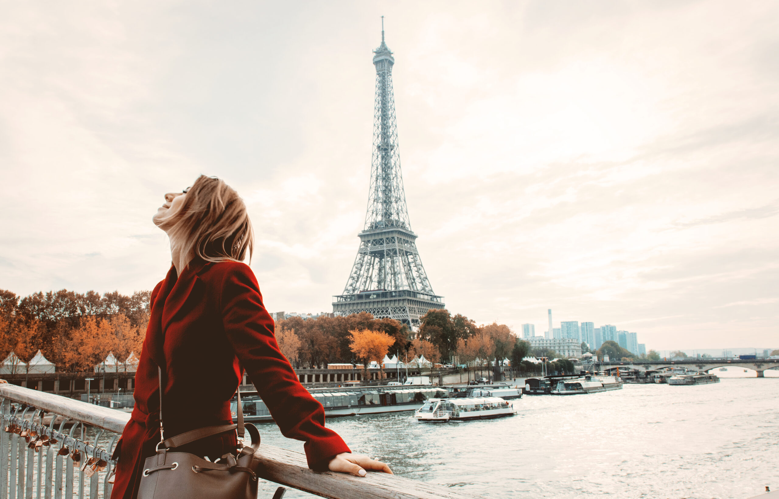 A woman in a red coat and a crossbody bag stands on the river Siene in fall, enjoying the weather and not worrying about what to wear in Paris in fall, the Eiffle tower behind her. 