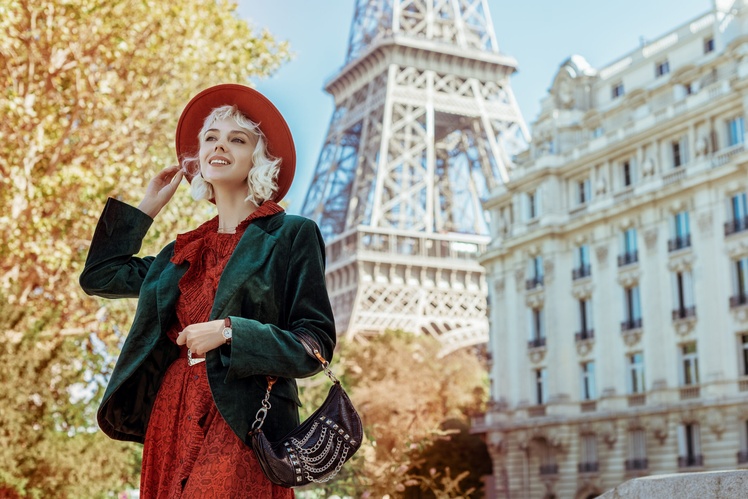 A woman in red dress and a green blazer and small clutch smiles off in the distance while the Eiffel Tower stands behind her. 