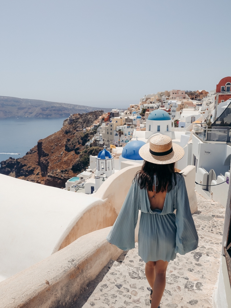 A woman navigates the stairs in Santorini in a straw hat and a long sleeved, open back, blue sundress. 