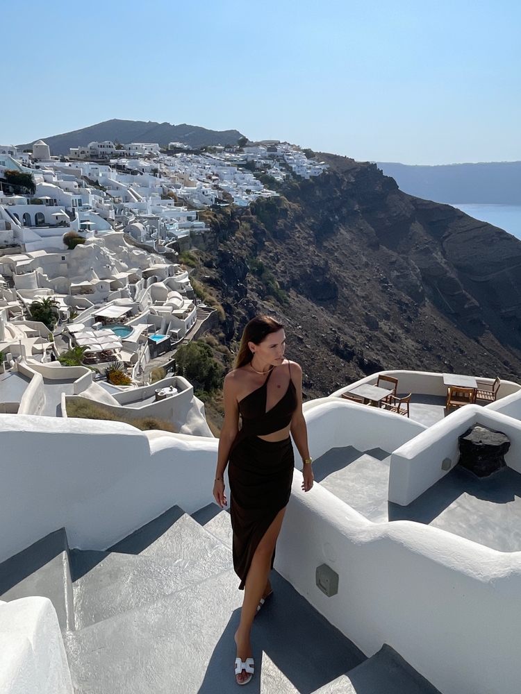 Ready to go out, this model looks out over the views of Santorini in a black dress that has cut outs. She pairs it with gold jewelry and white shoes. 