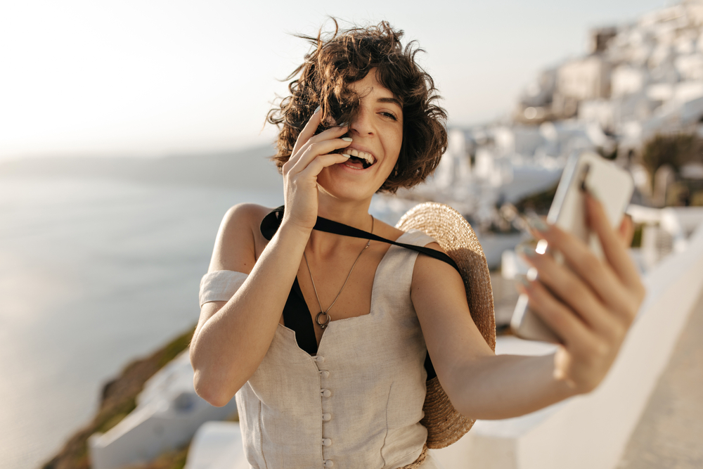 A woman poses for a selfie in her linen, button down top, straw hat, and wild curls on the coast. 