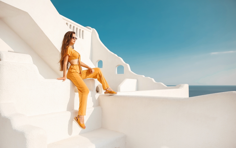 A woman in a bight yellow two piece set sits on a white wall, knowing what to wear in Santorini Greece, as her color is a splash of contrast in the setting. 