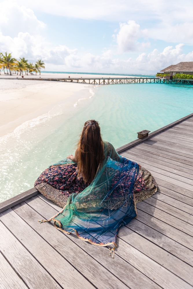 A woman with a flowy maxi dress and a colorful Shawl over it sits not he edge of a dock on the water. 