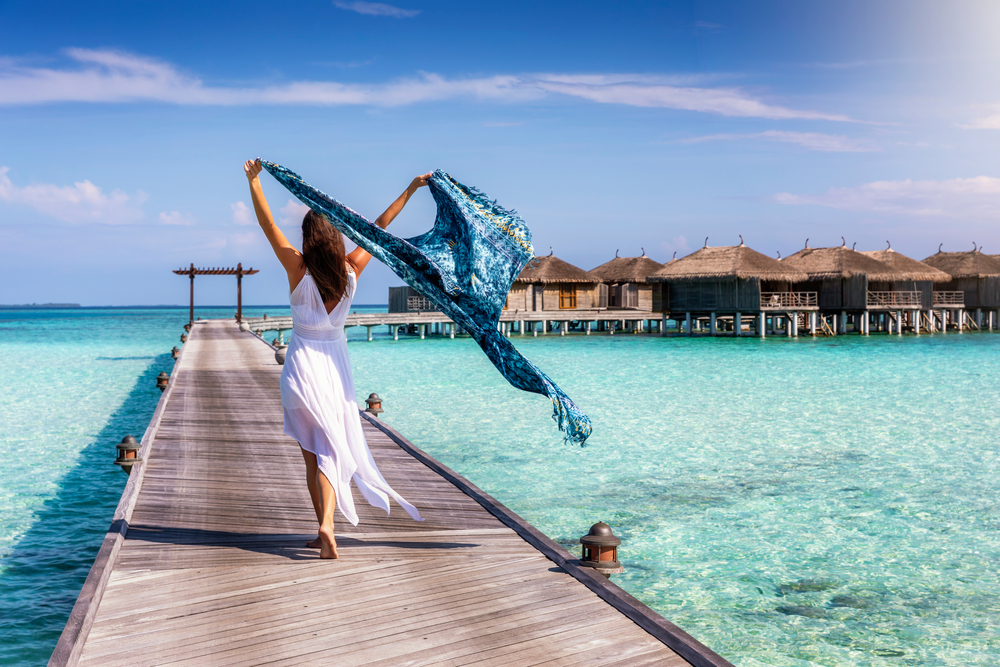 A woman in a flowy dress with a tropical shawl lets the shawl float in the wind, knowing what to wear in the Maldives, as she makes her way down the dock and to the bungalow resorts. 