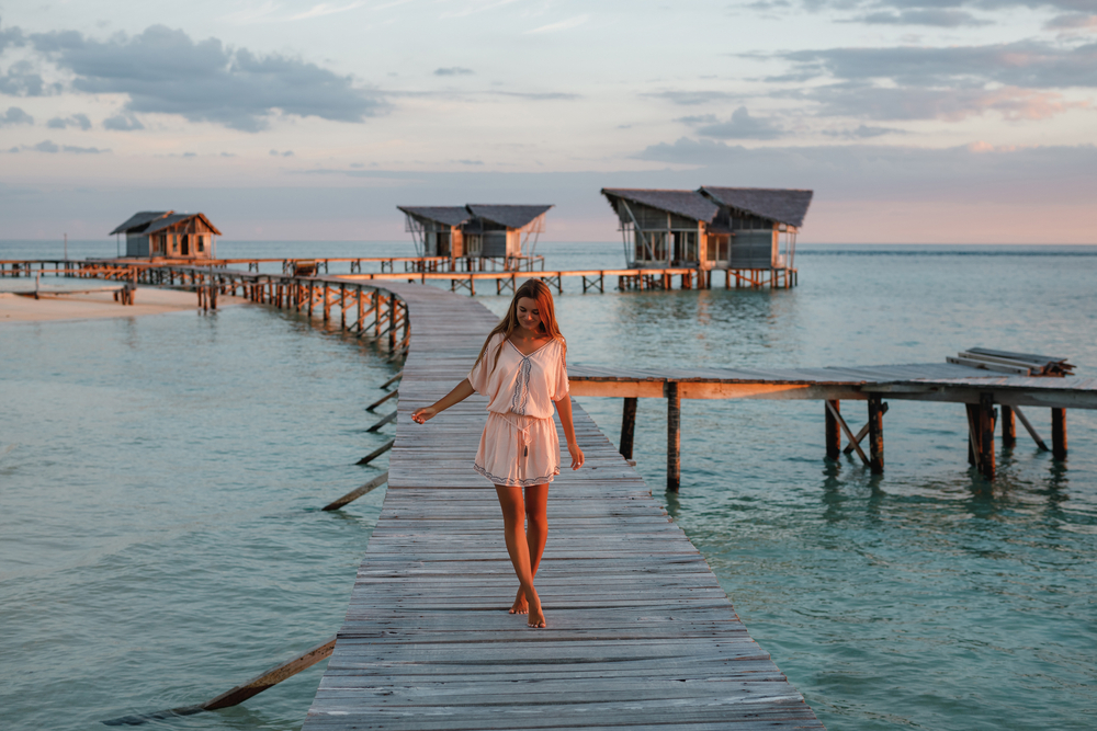 A woman tip toes down a wooden dock that has off ramps of bungalows: she wears a Boho romper and is barefoot during golden hour, knowing exactly what to wear in the Maldives. 