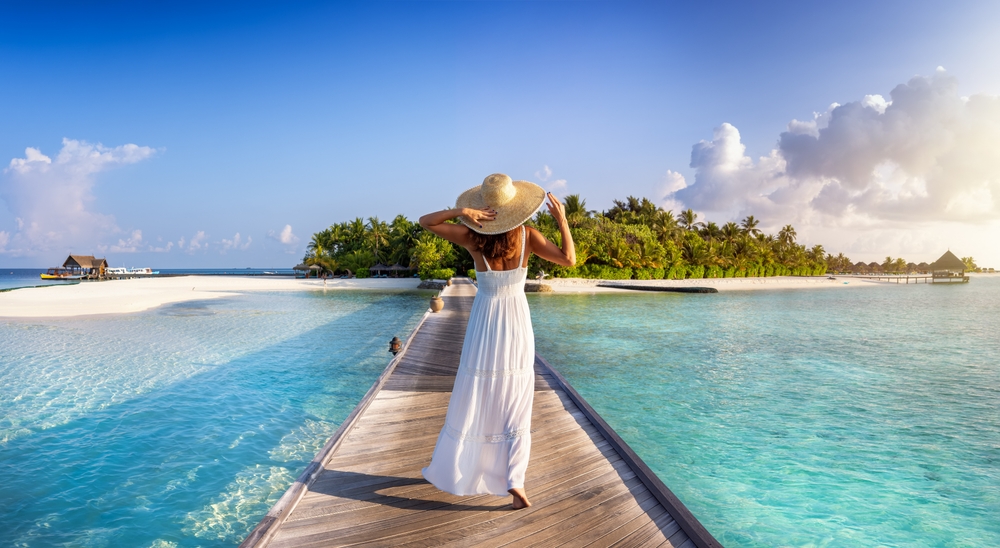 A woman in a long maxi dress, that is white, overlooks the island while on a dock, not worrying about what to wear in the Maldives, as she is comfortable and fashionable. 