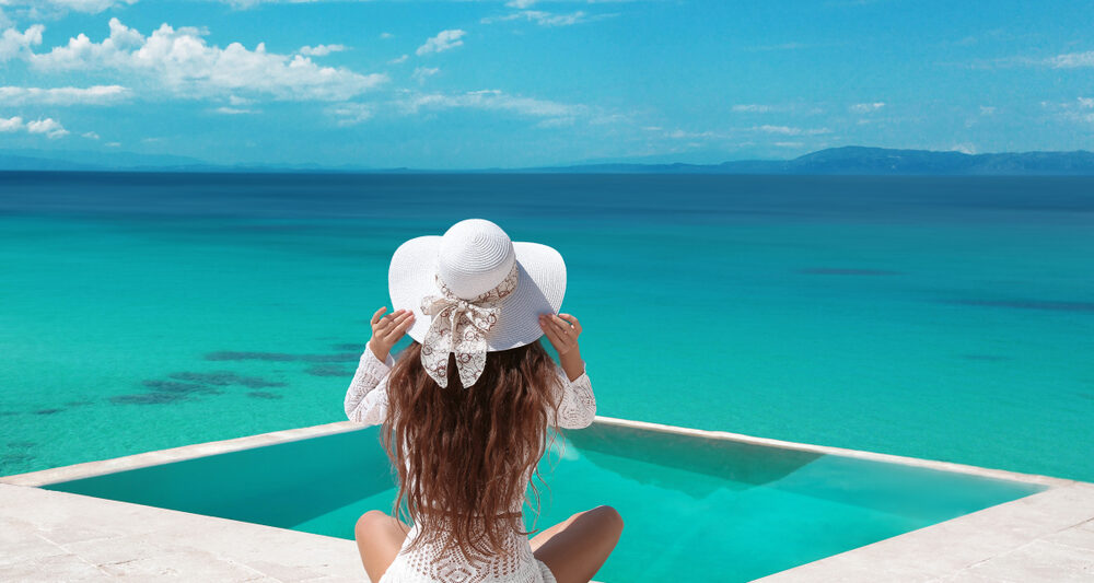 A woman sits on a dock in a white flowy dress, and a white straw hat, not worrying about what to wear in the Maldives as she is comfortable as she looks over the blue waters.