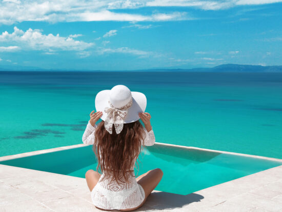 A woman sits on a dock in a white flowy dress, and a white straw hat, not worrying about what to wear in the Maldives as she is comfortable as she looks over the blue waters.