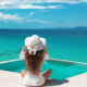 A woman sits on a dock in a white flowy dress, and a white straw hat, not worrying about what to wear in the Maldives as she is comfortable as she looks over the blue waters.