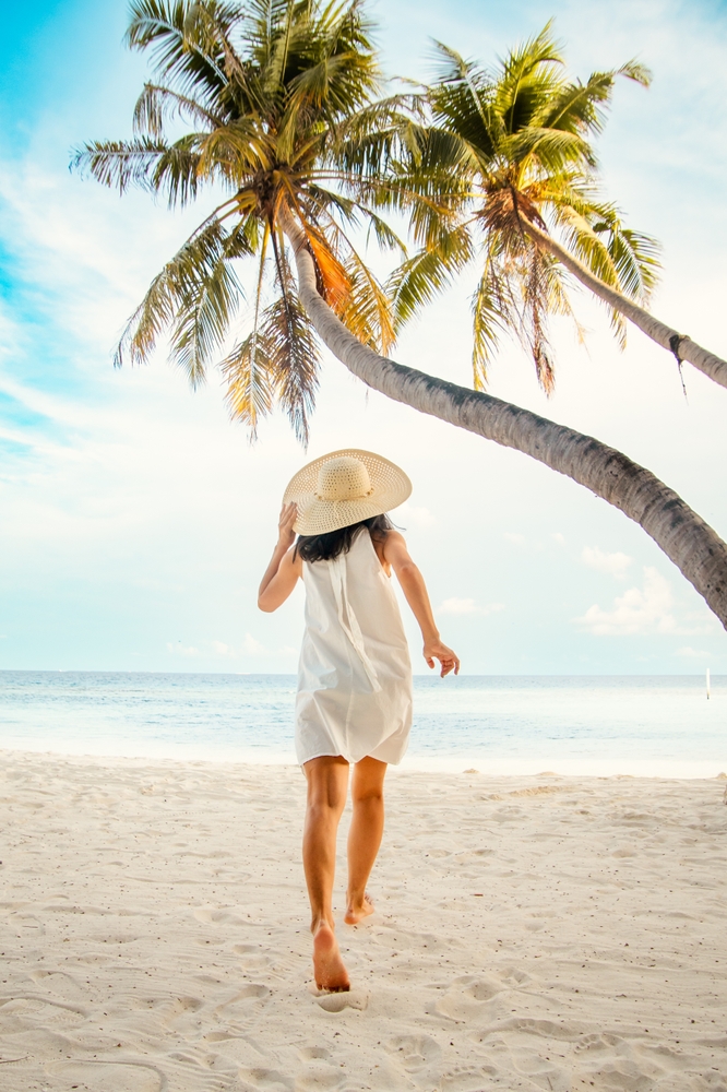 A woman in a soft linen t-shirt dress runs to the beach: she is barefoot and palm trees stretch tall above her. 