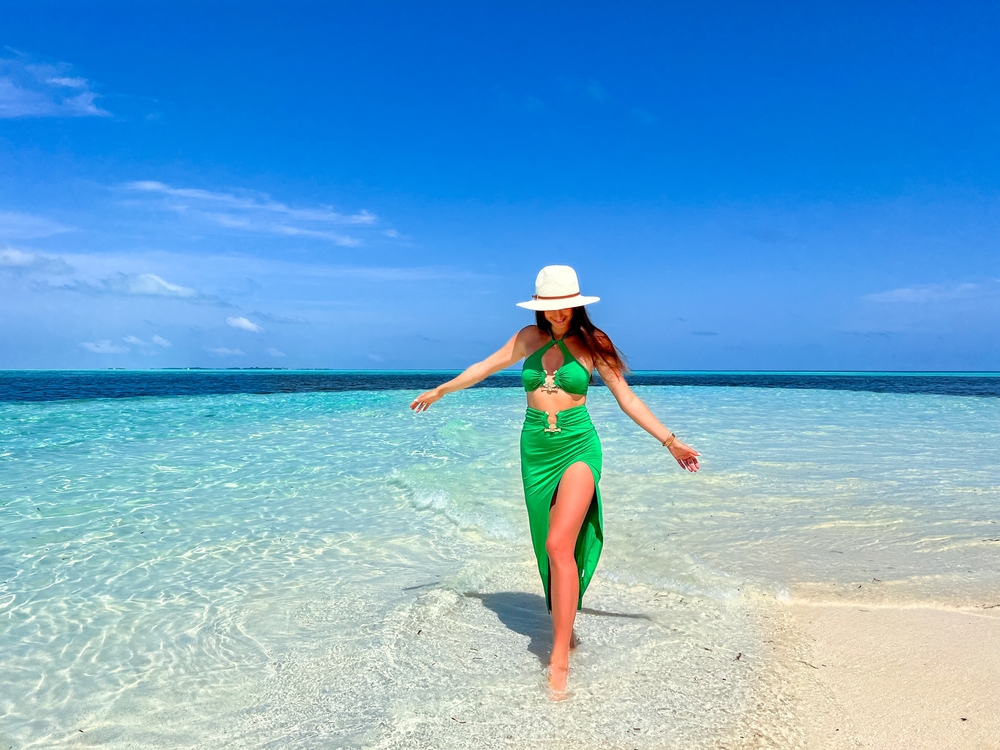 A woman with a two piece set, bright green in color, is comfortable as she walks through the clear water while wearing a sun hat: she knows exactly what to wear in the Maldives. 