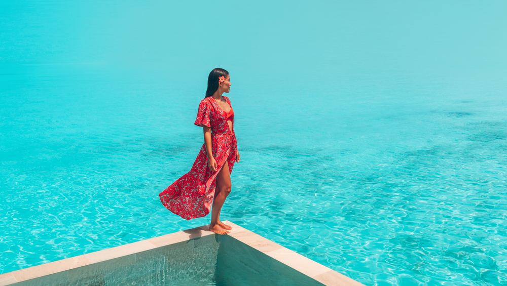 A woman in a red bikini with a tropical print/red cover up stands on the ledge of a resort and overlooks the water below. She has a red flower in her hair. 