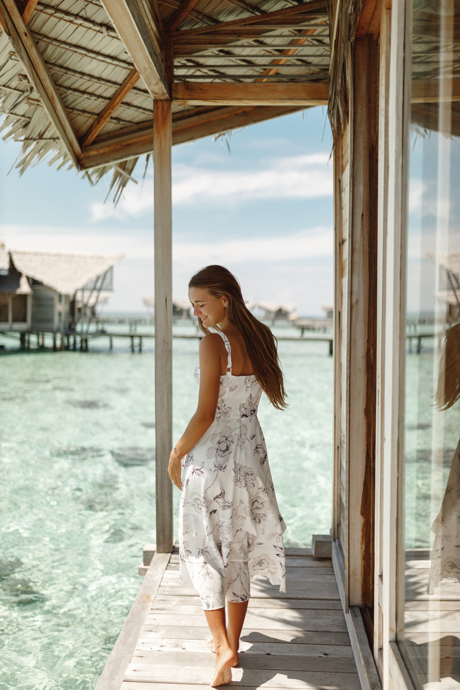 A woman with long hair knows exactly what to wear in the Maldives: her midi dress is white with tropical prints and its thin straps keeps her cool as she overlooks the edge of her resort room and into the water. 