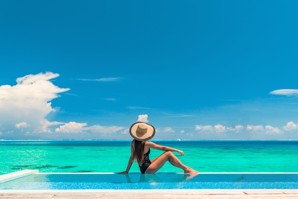 A woman in a black one piece swim suit with cut outs sits on the edge of a pool that also overlooks the water of the beach. 