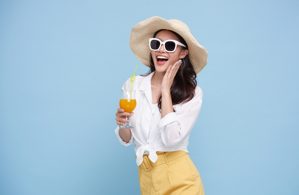 A young woman in a straw hat, sunglasses, a white button up and yellow shorts holds a drink and smiles. 