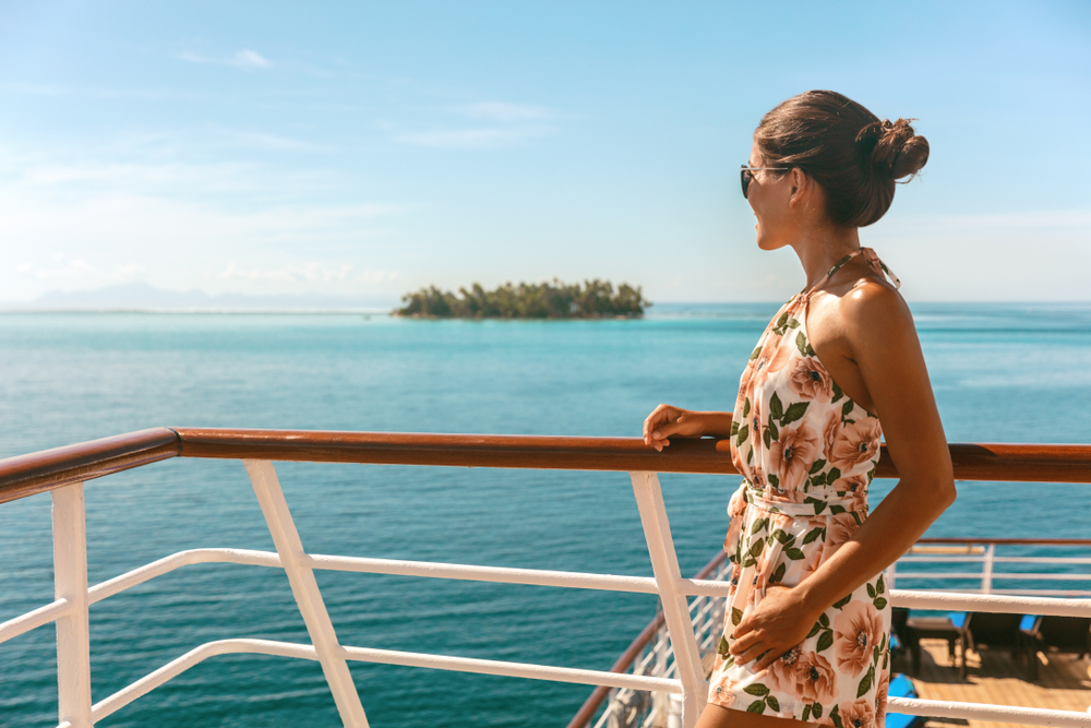 A woman on the edge of a boat in a floral sundress and sunglasses and a bun looks over the blue waters. 