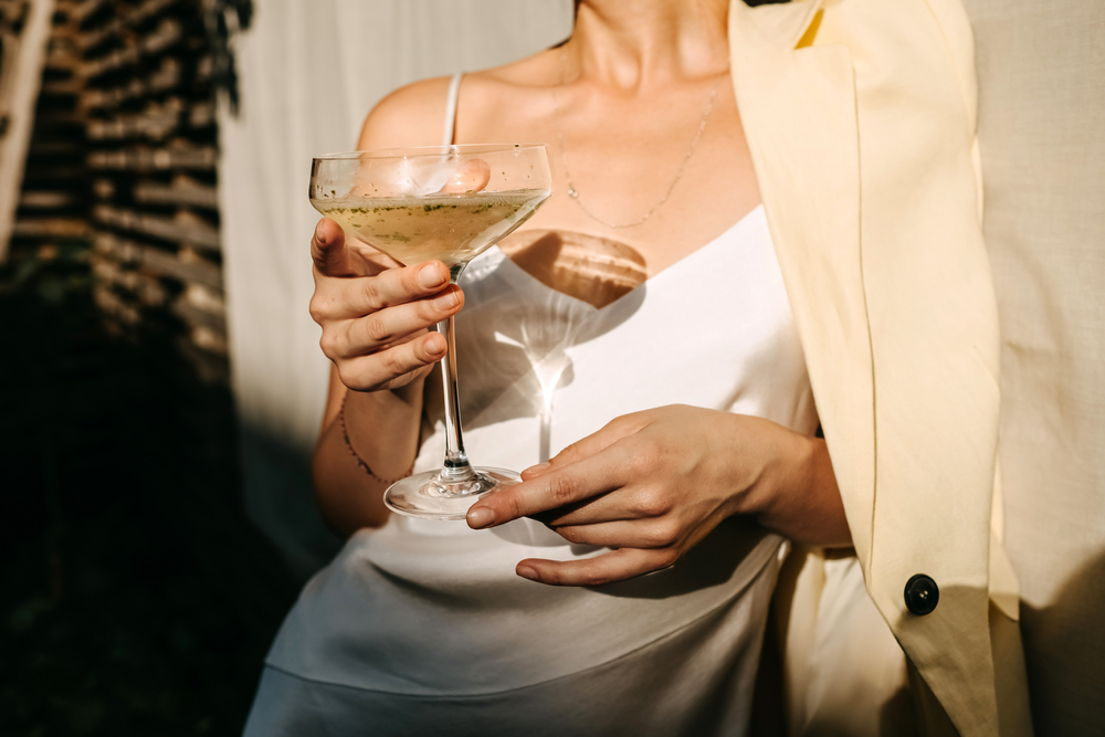 A woman holds a cocktail in a white, silky, formal gown, with a beige blazer thrown over one shoulder, which is perfect for what to wear on a cruise for cocktail parties! 