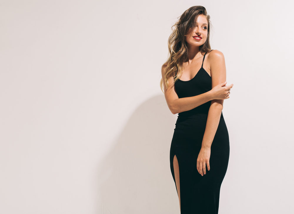 A woman poses in a spaghetti strap black dress with a slit up the side, exuding elegance with her red lipstick and curled hair. 