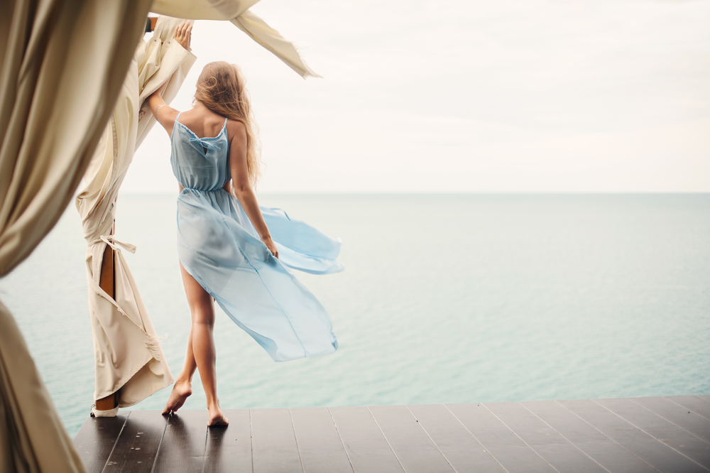 A woman in a blue, lengthy maxi dress that flows the the side in the wind stands on the edge of a boardwalk and looks at the water. These outfits are perfect for what to wear on a cruise because you can dress it up or dress it down. 