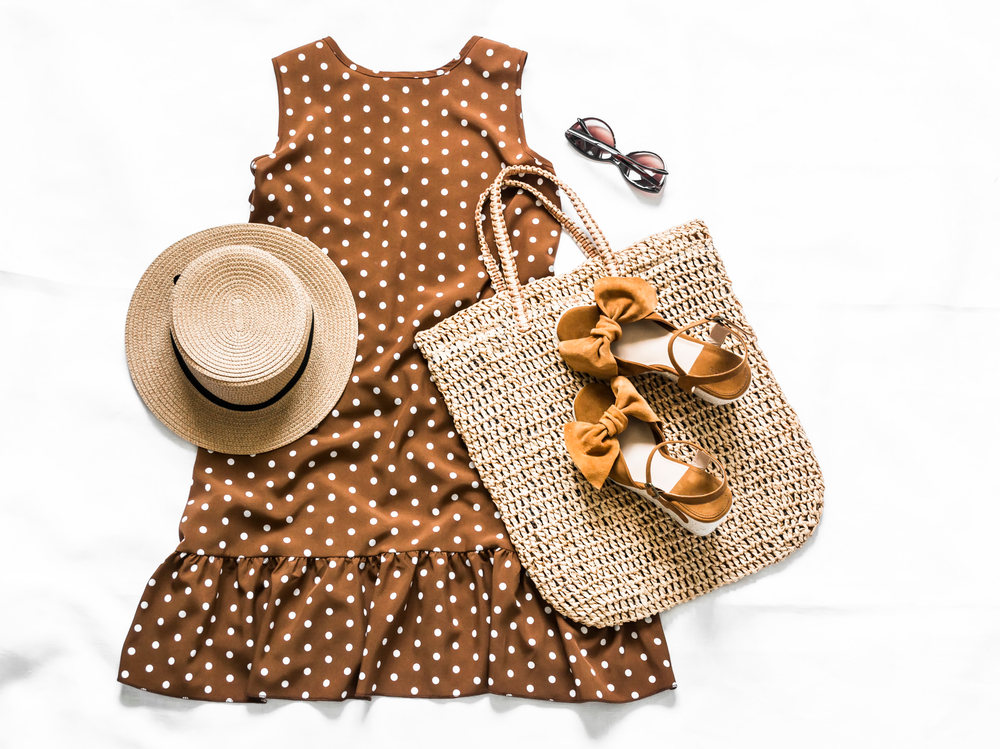 An outfit idea for a cruise includes a cute sundress that is brown with white polka dots, a straw hat, sunglasses, wedges, and a straw/knit tote bag. 