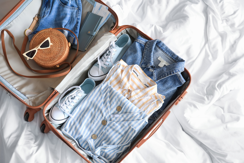 A packed suitcase is filled with linen clothing and jeans: button downs, sneakers, sunglasses and more. All in a blue hue! 