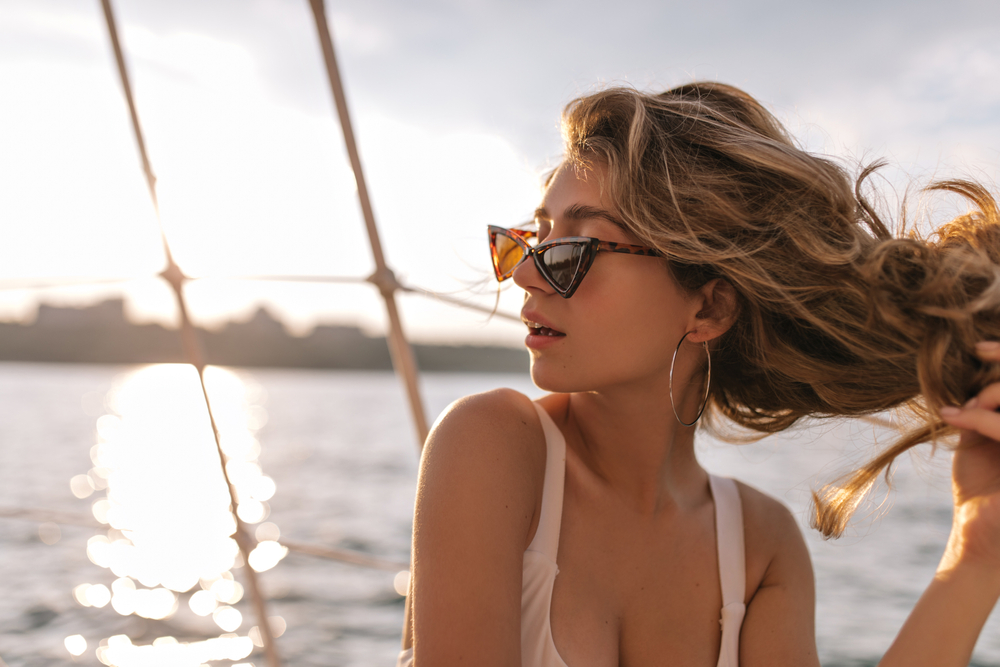 A woman pulls at her hair as she looks over her shoulder in a beige tank top and statement sunglasses on the edge of a boat, content in what to wear on a cruise during the sunset behind her. 