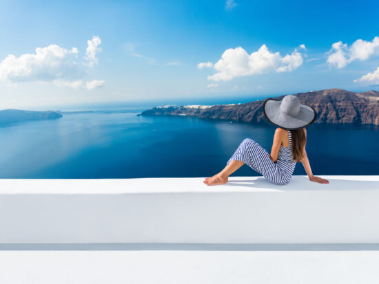 woman sitting in santorini greece wearing a blue and white striped outfit with a floppy hat overlooking the ocean