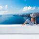 woman sitting in santorini greece wearing a blue and white striped outfit with a floppy hat overlooking the ocean