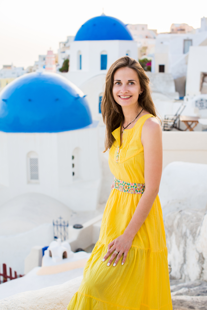 A woman smiles in Santorini in a yellow sundress, the color a bright splash in contrast to the blue and white buildings behind her. 