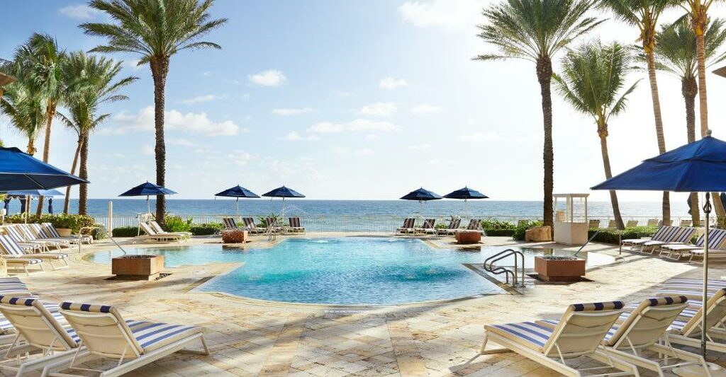 the oceanfront property with pool and palm tree and ocean. there are blue umbrellas around and green grass aera and tan tiles surrounding the pool.