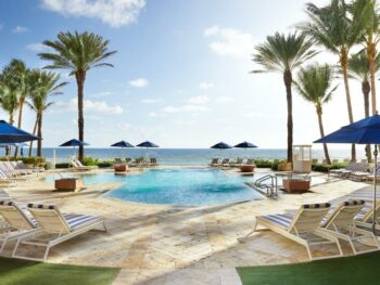 the oceanfront property with pool and palm tree and ocean. there are blue umbrellas around and green grass aera and tan tiles surrounding the pool.