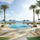 the oceanfront property with pool and palm tree and ocean. there are blue umbrellas around and green grass aera and tan tiles surrounding the pool.