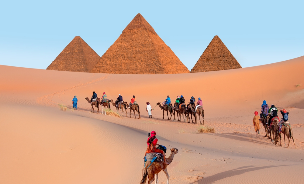 people ride camels through the desert in Egypt, there are pyramids in the background and a clear sky in egypt