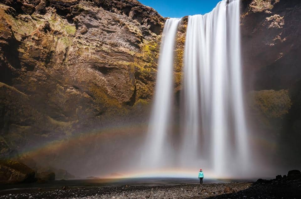 a large waterfall in iceland making a rainbow with someone standing at the bottom of it 