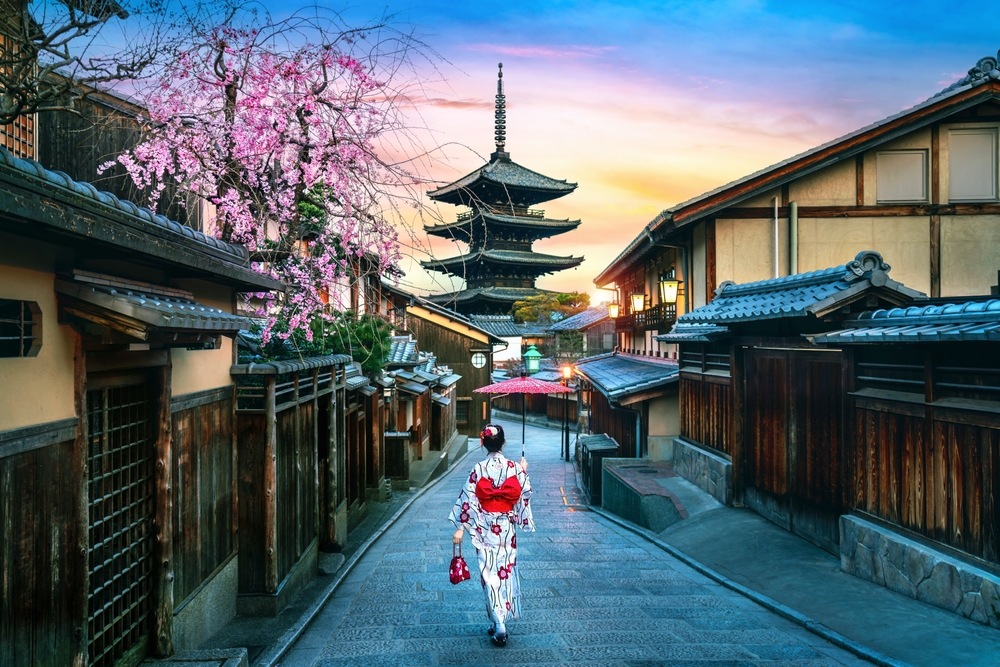 a woman is wearing a kimono in a street in japan, cheery blossoms are blooming on the left and a large structure can be seen in the background